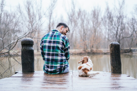 Smiling Hispanic Man Wearing Flannel With Cavalier King Charles Spaniel On A Wooden Dock In Lake