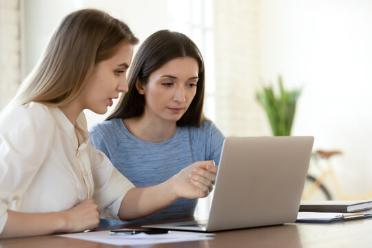 Focused young female leader helping newbie colleague with software applications, working on computer in office. Two serious busy teammates developing online marketing strategy or discussing project.