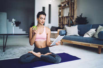 Naklejka na ściany i meble Slim fit girl enjoying fresh snack after active training workout in home interior using modern touch pad for watching online video,millennial yogi with green apple reading sportive network publication