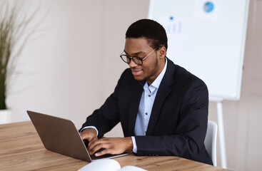 Young black manager smiling while working with laptop, office interior