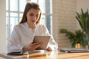 Smiling attractive young female doctor holding digital computer touchscreen tablet in hands, consulting patient online or web surfing information, working on modern tech gadget in clinic office.