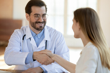 Fototapeta premium Smiling young handsome male gp doctor in eyeglasses shaking hands with happy female patient, welcoming at visit, making agreement or signing deal at consultation, getting acquainted in clinic office.