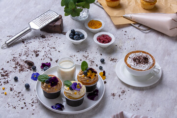 Gourmet cupcakes, cappuccino and chocolate chips. Cupcakes covered with edible flowers. A large piece of chocolate, raw cupcakes, a cup of coffee with cream and creamy, cappuccino. Dessert design