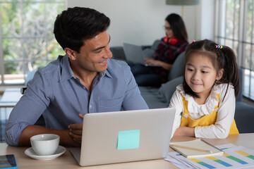 Mixed race family staying together, Caucasian father sitting and working at desk and teaching half-race little cute daughter white Asian mother using laptop computer on sofa. Idea for work at home - 414129709