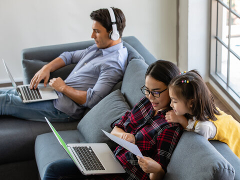 Multiracial Family, Father, Mother, And Little Daughter Stay Together At Home. Mom And Dad Working With Laptop Notebook And Teacing The Girl. Work From Home And New Normal Concept