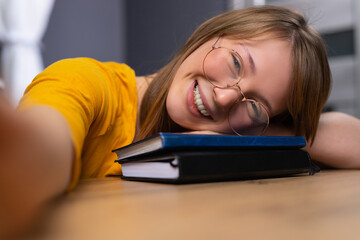 Young blonde, wearing yellow blouse and eyeglasses, taking a selfie, holding her head on the books. Student with black and blue books, smiling. Studying fun. Cute girl.