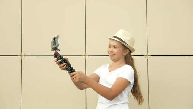 Free Girl blogger leads an online videoconference from city street. Fashionable girl in hat makes selfie outdoors. A teenager is photographed on smartphone using small tripod. Digital technologies