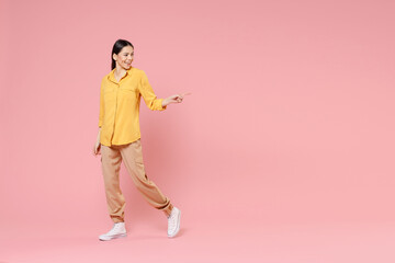 Full length of young smiling brunette positive attractive latin woman 20s in yellow casual shirt walk point index finger aside on workspace area isolated on pastel pink background studio portrait.