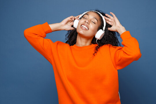 Smiling young african american woman in casual bright orange sweatshirt listening music with headphones dancing put hands on head keeping eyes closed isolated on blue color background studio portrait.