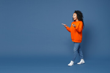 Full length side view of shocked young african american woman in casual orange sweatshirt standing pointing index fingers aside on mock up copy space isolated on blue color background studio portrait.