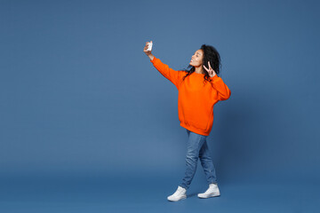 Full length of cheerful young african american woman in casual bright orange sweatshirt standing doing selfie shot on mobile phone showing victory sign isolated on blue background studio portrait.