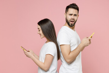 Young couple two friends man woman 20s in white blank design t-shirts hold in hand using mobile cell phone chatting typing sms standing back to back isolated on pastel pink background studio portrait.