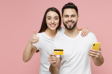 Young couple two friends man woman in white basic blank print design tshirts hold in hand using mobile cell phone credit bank card order home delivery in restaurant isolated on pastel pink background