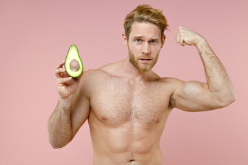 Strong bearded naked young man 20s perfect skin hold half of fresh avocado fruit showing biceps...