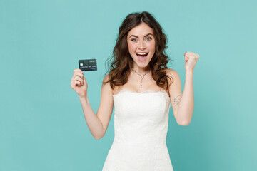 Excited bride young woman 20s in beautiful white wedding dress hold credit bank card doing winner gesture isolated on blue turquoise background studio portrait. Ceremony celebration party concept.