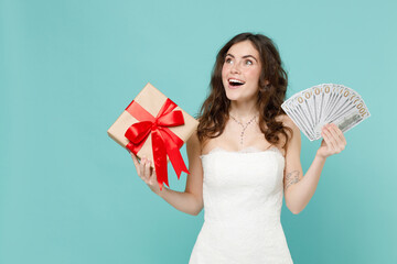 Shocked bride young woman in white wedding dress hold present box with gift ribbon bow fan of cash money in dollar banknotes isolated on blue turquoise background. Ceremony celebration party concept.
