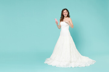 Full length excited bride young woman 20s in white wedding dress pointing index finger on mobile cell phone isolated on blue turquoise background studio portrait. Ceremony celebration party concept.