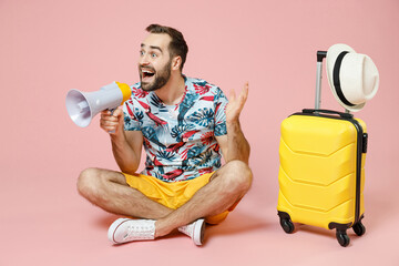 Full length of excited traveler tourist man in summer clothes sit on floor near suitcase screaming in megaphone isolated on pink background. Passenger traveling on weekend. Air flight journey concept.