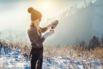 Woman backpacker traveler camper drinking hot tea and relaxing in winter forest, beautiful snow landscape on sunny day. Health care, authenticity, sense of balance.