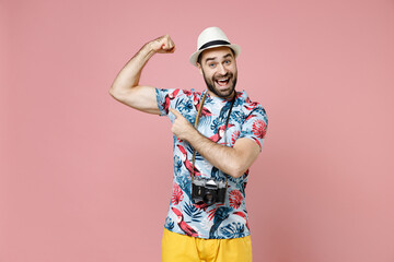 Excited young traveler tourist man in summer clothes hat with photo camera point index finger on biceps muscles isolated on pink background. Passenger traveling on weekend. Air flight journey concept.