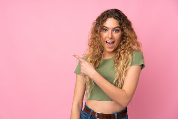 Young blonde woman with curly hair isolated on pink background surprised and pointing side