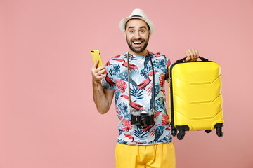 Excited young traveler tourist man in summer clothes hat hold suitcase using mobile phone booking...