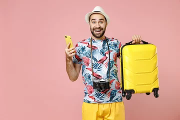 Foto op Canvas Excited young traveler tourist man in summer clothes hat hold suitcase using mobile phone booking taxi hotel isolated on pink background. Passenger traveling on weekends. Air flight journey concept. © ViDi Studio