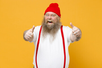 Fototapeta na wymiar Fat pudge obese chubby overweight tattooed blue-eyed bearded excited happy man 30s has big belly in white t-shirt red hat suspenders show thumbs up like gesture isolated on yellow background studio