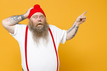 Fototapeta na wymiar Fat pudge obese chubby overweight tattooed bearded shocked man has big belly in white t-shirt red hat suspenders point index finger aside on workspace hold head isolated on yellow background studio.