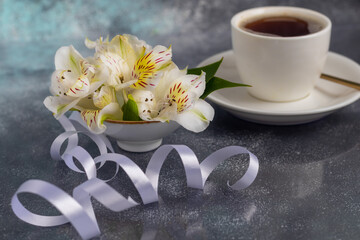 Obraz na płótnie Canvas White cup with coffee on a gray background. A bouquet of orchids entwined with a ribbon in the background. Banners, congratulations on the holiday.