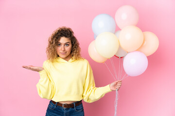 Young blonde woman with curly hair catching many balloons isolated on pink background having doubts with confuse face expression