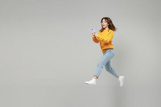 Full length of young fun smiling caucasian student woman 20s wearing knitted yellow sweater jumping high hold mobile cell phone chatting typing sms isolated on grey color background studio portrait..