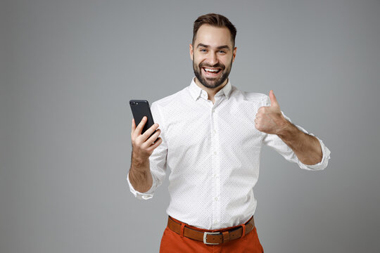 Cheerful young bearded business man in classic white shirt using mobile cell phone typing sms message showing thumb up isolated on grey background studio portrait. Achievement career wealth concept.