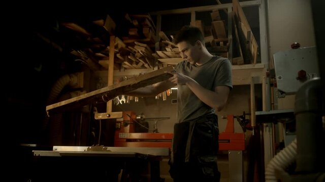 Young carpenter looking carefully at the piece of plank and checking it's width for making a beautiful piece of furniture and looking at it with love in his workshop.