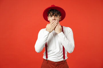 Young spanish latinos woondered surprised shocked man 20s in casual hat white shirt trousers with suspenders looking camera covering mouth with hands isolated on brightred background studio portrait.