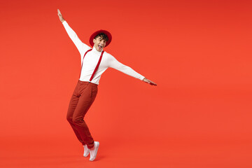 Full length of young spanish latinos surprised stylish man 20s in hat white shirt trousers with suspenders leaning over with outstratched hands look aside isolated on red background studio portrait.