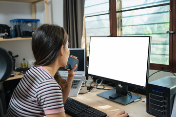 Young asian woman analyzing with holding mobile phone at working table with empty monitor in the house