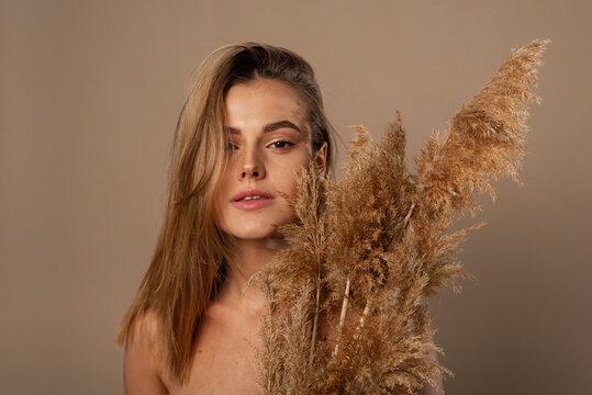 Close Up Beauty Portrait Of An Attractive Young Topless Woman With Brown Hair Isolated Over Beige Background, Holding Dried Herb. Skin Care Concept