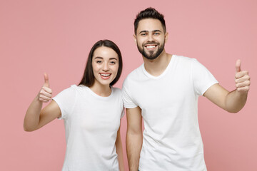 Young cheerful couple two friends bearded man brunette woman in white basic blank print design t-shirts smiling showing thumbs up like gesture isolated on pastel pink color background studio portrait.