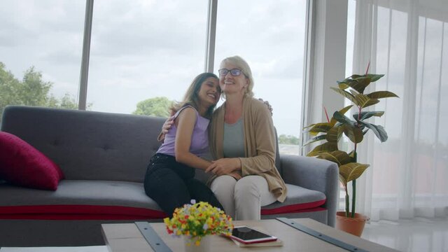 Wide shot - Adopted Asian teen daughter hugs her Caucasian stepmother after announcing good news, then smiles happily. Love and sharing time of diverse cultural family.