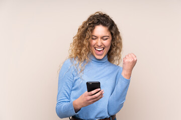 Naklejka premium Young blonde woman with curly hair wearing a turtleneck sweater isolated on beige background with phone in victory position