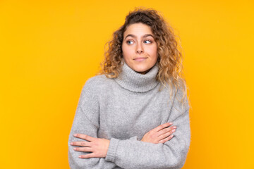 Fototapeta na wymiar Young blonde woman with curly hair wearing a turtleneck sweater isolated on yellow background making doubts gesture while lifting the shoulders