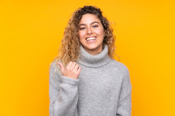 Young blonde woman with curly hair wearing a turtleneck sweater isolated on yellow background pointing to the side to present a product