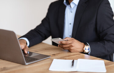 Unrecognizable black manager holding smartphone, using laptop