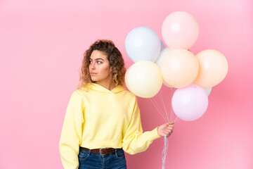 Young blonde woman with curly hair catching many balloons isolated on pink background looking side
