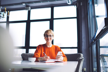Positive female freelancer taking notes while browsing smartphone in cafe