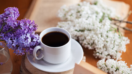 Spring mood, a cup of aromatic espresso coffee on a wooden table, branches of blooming lilac and a book