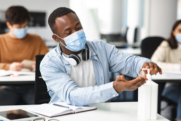 Black male student applying antibacterial sanitizer on hands - obrazy, fototapety, plakaty