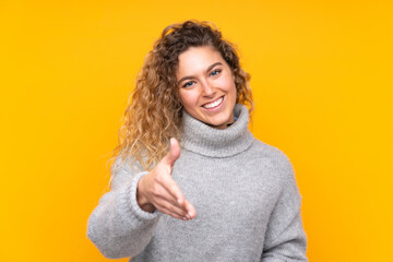 Young blonde woman with curly hair wearing a turtleneck sweater isolated on yellow background shaking hands for closing a good deal