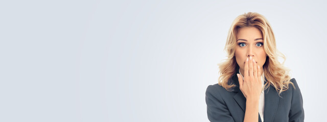 Shocked businesswoman in grey confident suit, with wide open eyes, covering mouth by her hand,...